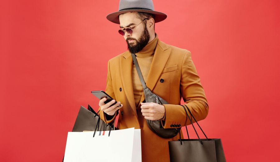 Foto von Max Fischer von Pexels, das eine Person mit Hut, Sonnenbrille, Pulli und Sakko zeigt, die Einkaufstüten trägt und auf ihr Smartphone schaut.