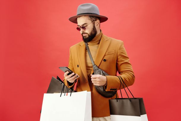 Photo by Max Fischer from Pexels, showing a person wearing a hat, sunglasses, sweater, and jacket, carrying shopping bags and looking at their smartphone.