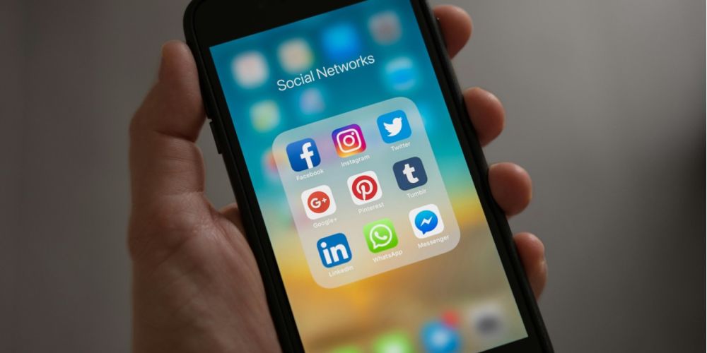 Eine Hand die ein altes iPhone hält, das Display zeigt den Home Bildschirm mit dem Ordner Social Networks geöffnet, darin befinden sich App Icons von Facebook, Instagram und mehr.