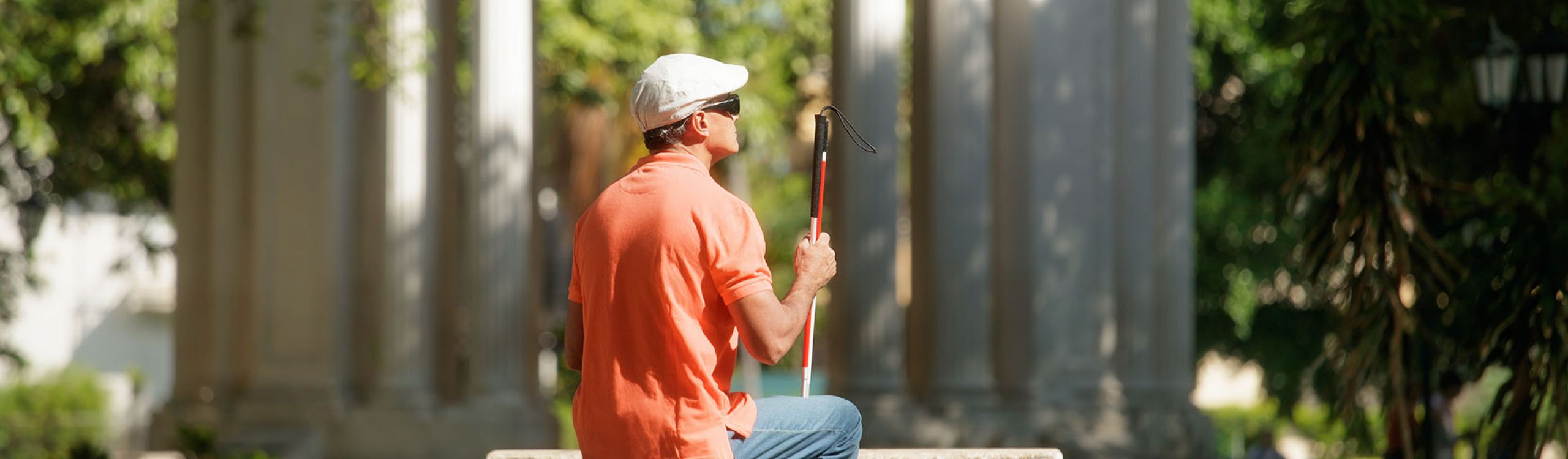 A blind man wearing blue jeans, a red polo shirt and a white cap sits on a stone bench with his long cane.